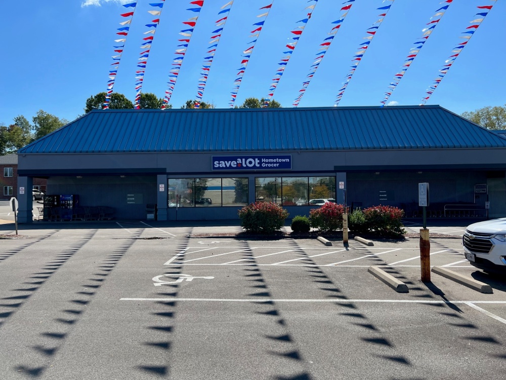 A grocery store with a blue roof and a "Save a Lot" sign displays its grocery store license proudly. Colorful triangular flags are strung above the building, and the parking lot in front has marked spaces and a few parked cars.